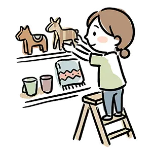 脚立に登って棚におしゃれな木彫りの雑貨やカップを陳列・整頓する女性ショップ店員